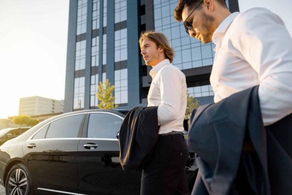 Two businessmen get in luxury car near hotel or office building on sunset, going to drive after work. Concept of transportation and business lifestyle